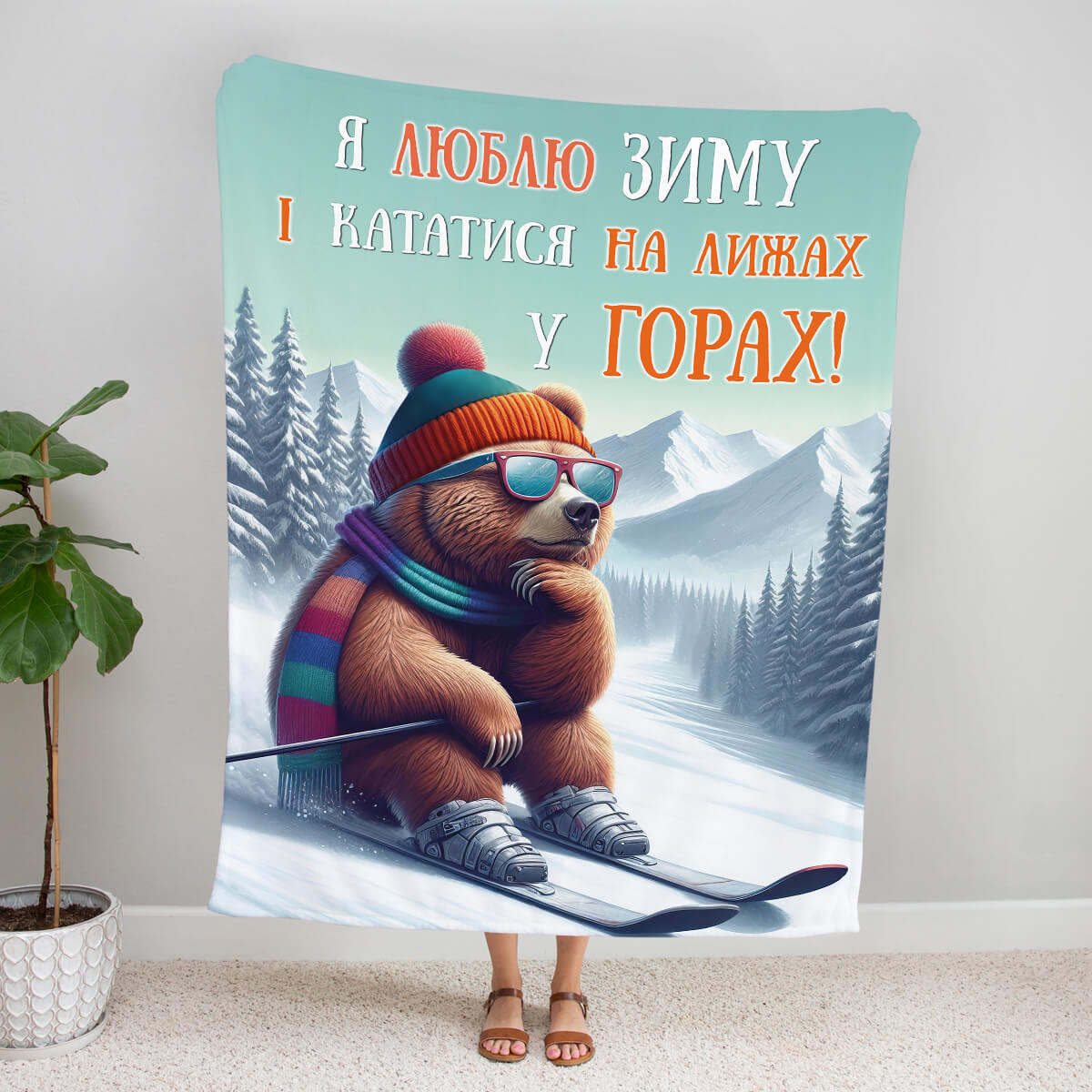 Плед із бурим ведмедем у шапці з помпоном, окулярах і шарфі на лижах серед засніжених гір